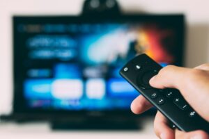 A hand holding a streaming remote control pointed at a connected TV, illustrating how to buy television advertising through CTV platforms.