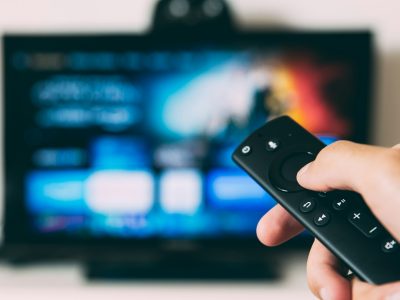 A hand holding a streaming remote control pointed at a connected TV, illustrating how to buy television advertising through CTV platforms.